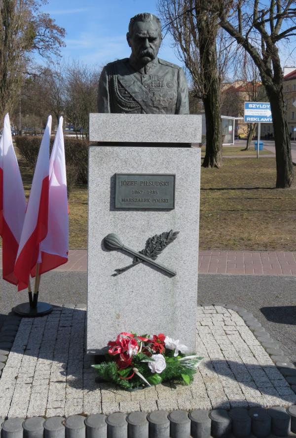 Popiersie mężczyzny z wąsami w mundurze z odznaczeniami na jasnym obelisku z tablicą Józef Piłsudski Marszałek Polski.