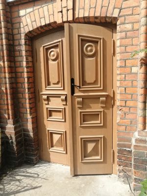 Dwuskrzydłowe drzwi z jednym skrzydłem uchylonym. Na drzwiach rzeźbione w drewnie dekoracje w stylu z przełomu XIX i XX w.