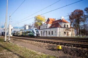Ozdobny, beżowy budynek zabytkowy z dachem z czerwonej dachówki. Przed nim biało-zielono-żółty pociąg Kolei Mazowieckich.