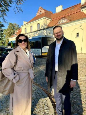 Kobieta w beżowym płaszczu i okularach oraz mężczyzna w czarnym płaszczu i okularach na tle białego autobusu i zabytkowego...