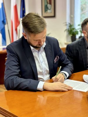 Mężczyzna w marynarce, siedzący przy stole podpisuje dokument.