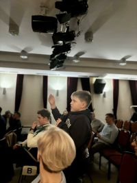 Chłopiec w czarnej bluzie z kapturem unosi prawą rękę do góry, a w lewej trzyma mikrofon.
