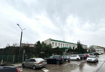 Kilkanaście różnokolorowych samochodów na parkingu prze białym prostokątnym budynkiem.