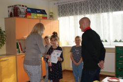 Kobieta wręcza dziewczynce dyplom i medal na biało-czerwonej wstążce.