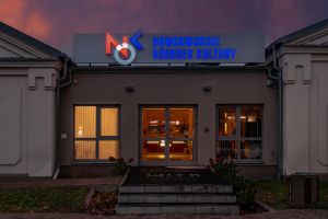 Nad szklanym wejściem do budynku kolorowy neon Nowodworski Ośrodek Kultury z logo.