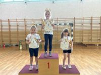 Trzy dziewczynki w koronach na głowie stoją na podium trzymając w rękach puchary i dyplomy.