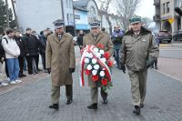 Trzech mężczyzn w mundurach wojskowych. Mężczyzna w środku trzyma wiązankę kwiatów.