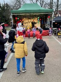 Osoba w stroju niedźwiedzia, w długiej czapce, dwie kobiety i i ośmioro dzieci tańczą w kółeczku przed sceną.