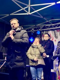 Za mężczyzną mówiącym do mikrofonu na scenie stoją dwie kobiety.