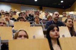 Trzy kobiety i kilkunastu mężczyzn siedzących na krzesłach za ławkami umieszczonymi kaskadowo na sali wykładowej.
