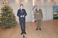 Mężczyzna w okularach ubrany w garnitur mówi do mikrofonu na statywie. Obok stoi Mężczyzna w mundurze wojskowym.