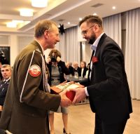 Mężczyzna w mundurze wojskowym i mężczyzna w garniturze stoją naprzeciwko siebie trzymając wspólnie w dłoniach pudełko.