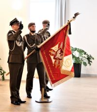 Trzech mężczyzn w mundurach wojskowych: jeden trzyma pochylony do przodu sztandar na drzewcu, dwóch po jego bokach salutuje.