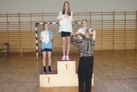 Trzy dziewczynki w koronach na głowach i z medalami na szyjach stoją na podium trzymając w dłoniach puchary i dyplomy.