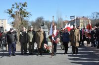 Mężczyźni w mundurach wojskowych i chłopiec ze sztandarem przed pomnikiem Marszałka Józefa Piłsudskiego.