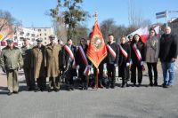 Młodzież w szarfach ze sztandarem i mężczyźni w mundurach wojskowych przed pomnikiem Marszałka Józefa Piłsudskiego.