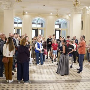 Kilkadziesiąt osób stojących w zabytkowym holu, patrzących na ozdobny sufit i żyrandole.