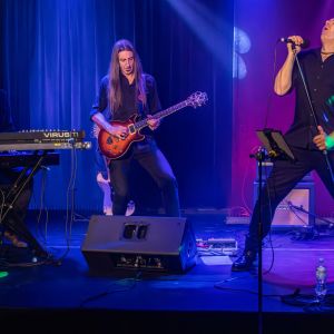 Mężczyzna grający na keyboardzie i mężczyzna grający na gitarze. Obok nich mężczyzna z odchyloną do góry głową trzyma w...