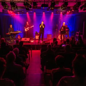 Pięciu mężczyzn na podświetlonej scenie, grających na keyboardzie, perkusji, dwóch na gitarach i jeden trzymający w...