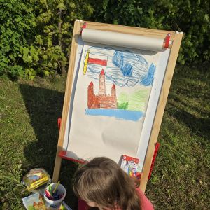 Dziewczynka siedząca obok rysunku, zamku i biało-czerwonej flagi, umieszczonego na sztaludze.