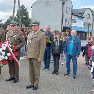 Trzech mężczyzn w mundurach i czapkach wojskowych stojących obok siebie. Mężczyzna w środku trzyma w rękach wiązankę kwiatów.