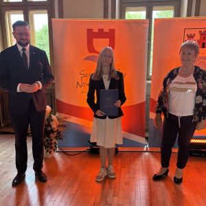 Dziewczynka z teczką trzymaną w dłoniach stoi pomiędzy mężczyzną i kobietą.