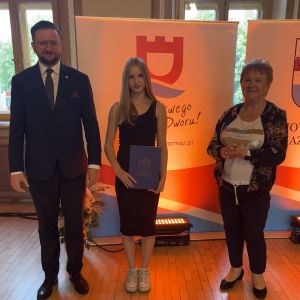Dziewczynka z teczką trzymaną w dłoniach stoi pomiędzy mężczyzną i kobietą.