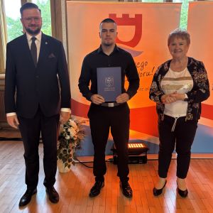 Chłopiec z teczką trzymaną w dłoniach stoi pomiędzy mężczyzną i kobietą.