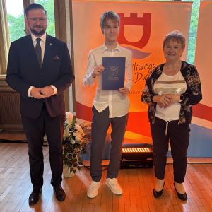Chłopiec z teczką trzymaną w dłoniach stoi pomiędzy mężczyzną i kobietą.