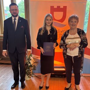 Dziewczynka z teczką trzymaną w dłoniach stoi pomiędzy mężczyzną i kobietą.