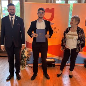 Chłopiec z teczką trzymaną w dłoniach stoi pomiędzy mężczyzną i kobietą.