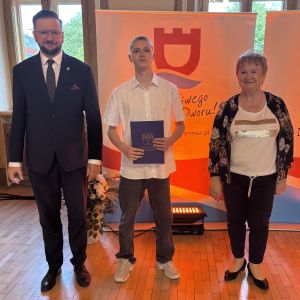Chłopiec z teczką trzymaną w dłoniach stoi pomiędzy mężczyzną i kobietą.