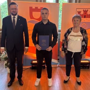 Chłopiec z teczką trzymaną w dłoniach stoi pomiędzy mężczyzną i kobietą.