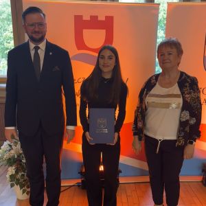 Dziewczynka z teczką trzymaną w dłoniach stoi pomiędzy mężczyzną i kobietą.