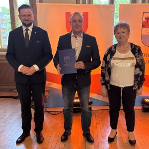 Mężczyzna z teczką trzymaną w dłoniach stoi pomiędzy mężczyzną i kobietą.