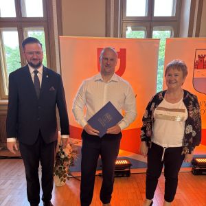Mężczyzna z teczką trzymaną w dłoniach stoi pomiędzy mężczyzną i kobietą.