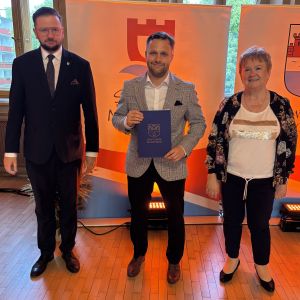 Mężczyzna z teczką trzymaną w dłoniach stoi pomiędzy mężczyzną i kobietą.