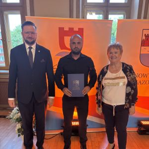 Mężczyzna z teczką trzymaną w dłoniach stoi pomiędzy mężczyzną i kobietą.