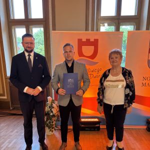 Mężczyzna z teczką trzymaną w dłoniach stoi pomiędzy mężczyzną i kobietą.