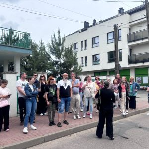 Grupa kobiet i mężczyzn stojących na chodniku patrzących w stronę kobiety stojącej przed nimi.