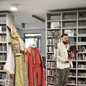 Mężczyzna trzymający bukiet róż w rękach stoi obok szat liturgicznych na stojakach.