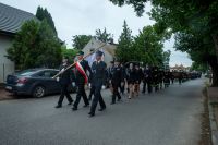Przemarsz pocztu sztandarowego Ochotniczej Straży Pożarnej z grupą kilkudziesięciu kobiet i mężczyzn w mundurach strażackich.