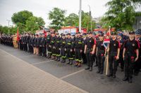 Kilka kobiet i kilkudziesięciu mężczyzn w mundurach strażackich stojących na placu na tle wozów strażackich.