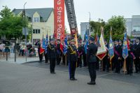 Trzech mężczyzn w mundurach strażackich zwróconych do siebie na tle dziesięciu pocztów sztandarowych.