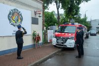 Trzech mężczyzn w mundurach strażackich salutujących przed tablicą, pod którą stoi wieniec z biało-czerwonych kwiatów.