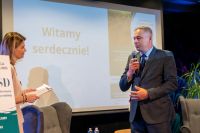Mężczyzna w garniturze mówi do mikrofonu trzymanego prawą ręką.