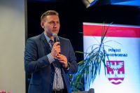 Mężczyzna w garniturze mówi do mikrofonu trzymanego prawą ręką.