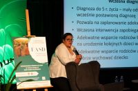 Kobieta w okularach oparta rękami o fotel mówi do mikrofonu trzymanego w prawej dłoni.