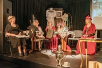 Cztery kobiety w ozdobnych sukniach siedzące na krzesłach na scenie.