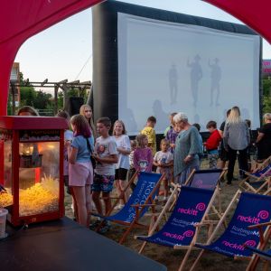 Grupa dzieci i czworo dorosłych przy pompowanym ekranie kinowym. Przed nimi leżaki plażowe i stoisko z popcornem.